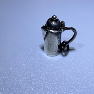 Può includere: Un piccolo ciondolo color argento a forma di caffettiera. Il ciondolo ha un manico, un coperchio e un beccuccio. Il ciondolo a forma di caffettiera è probabilmente realizzato in argento sterling ed è una rappresentazione in miniatura di una caffettiera classica.