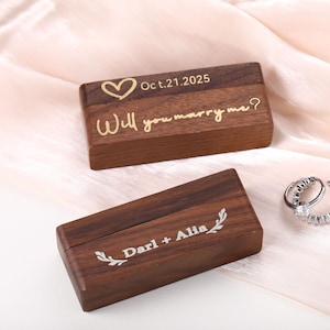 May include: Two wooden ring boxes with engraved text. One box reads "Will you marry me?" with a heart and "Oct.21.2025". The other box reads "Darl + Alia" with a leaf design. A silver ring set with clear stones is also present.