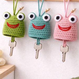 Könnte beinhalten: Drei handgefertigte Häkelschlüsselanhänger in Froschform in Grün, Türkis und Rosa. Jeder Frosch hat weiße Augen mit schwarzen Pupillen, einen roten, lächelnden Mund und einen Schlüssel, der unten heraushängt. Sie hängen an einer Holzleiste.