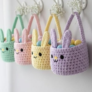 May include: Four pastel-colored crochet bunny baskets hanging on a white wall. Each basket has a handle, bunny ears, and a cute face. The baskets are filled with colorful Easter eggs and are decorated with small white flowers.
