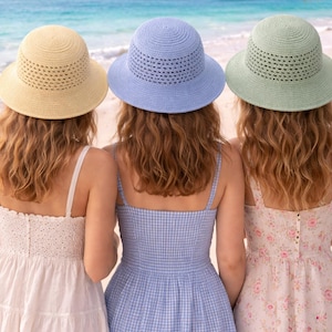 May include: Three women wearing summer hats in yellow, blue, and green stand on a beach. The hats have a woven design. They are wearing dresses in white, blue gingham, and floral patterns, with the ocean in the background.