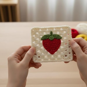 Può includere: Un sottobicchiere quadrato all'uncinetto color crema con una fragola rossa e foglie verdi. Il sottobicchiere è tenuto tra due mani, evidenziando i dettagli fatti a mano e il design traforato. La fragola è centrata sul sottobicchiere.