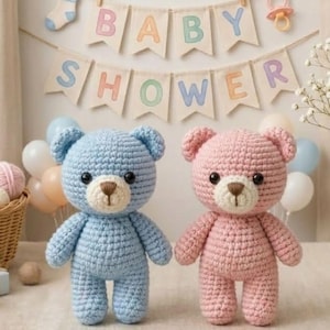 Puede incluir: Dos osos de peluche de ganchillo hechos a mano, uno azul claro y otro rosa, se encuentran frente a una pancarta que dice "BABY SHOWER". Los osos tienen narices marrones y ojos negros. Una cesta de hilo y globos están en el fondo.