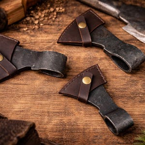 Könnte beinhalten: Nahaufnahme von drei kleinen, dunklen Metallbeilen mit braunen Lederscheiden und Messingknopfverschlüssen. Die Beile sind auf einer Holzoberfläche angeordnet, mit einer größeren Axt und anderen Werkzeugen im Hintergrund.