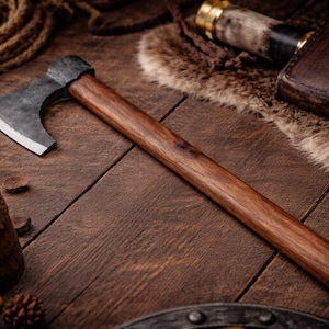 May include: A handcrafted axe with a dark metal head and a long, polished wooden handle. The axe is displayed on a wooden surface with other rustic items, including a leather-bound container and pine cones. The axe's blade is sharp and ready for use.