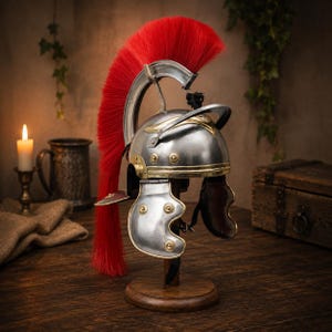 Puede incluir: Un casco romano plateado con detalles en latón y una pluma roja vibrante, exhibido en un soporte de madera. El casco presenta intrincados trabajos en metal y protectores de mejillas, evocando el equipo militar histórico. El fondo incluye una vela y un cofre de madera.