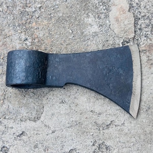 Könnte beinhalten: Ein schwarzer Axtkopf mit gebogener Klinge und zylindrischem Auge für den Stiel. Der Axtkopf besteht aus dunklem Metall und hat eine leicht strukturierte Oberfläche. Die Klinge ist silberfarben und scharf.