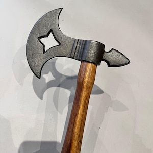 May include: A hand axe featuring a dark, textured metal head with a star-shaped cutout in the blade and a pointed poll. The axe head is securely attached to a smooth, polished wooden handle with a warm, reddish-brown grain pattern.