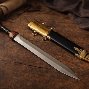Puede incluir: Una espada gladius romana con empuñadura de madera y hoja de acero, junto a su vaina de cuero negro con detalles dorados. La espada y la vaina están sobre una superficie de madera oscura.