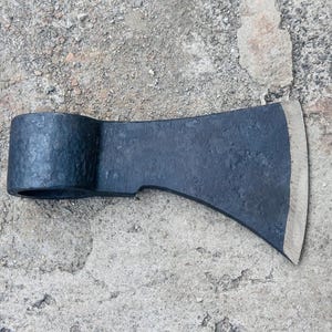 May include: A dark gray axe head with a curved blade and a cylindrical eye for the handle. The axe head is lying on a rough, gray surface. The blade's edge is silver.