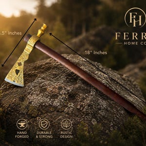 May include: A hand-forged tomahawk with a dark wooden handle and a decorative gold-coloured head. The head measures 21.59 cm, and the overall length is 45.72 cm. The tomahawk is displayed on a rock with a blurred forest background.