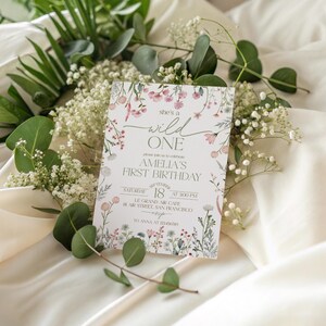 Puede incluir: Una tarjeta de invitaci&oacute;n con tem&aacute;tica floral para un primer cumplea&ntilde;os, con el texto "she's a wild ONE". La tarjeta est&aacute; rodeada de vegetaci&oacute;n y flores blancas, sobre una tela color crema.
