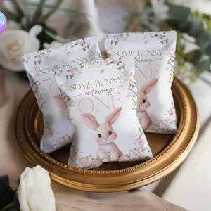 May include: Three white favor bags with a floral design and a bunny illustration. The bags are on a gold-colored tray. Text on the bags reads "SOME BUNNY is turning ONE."