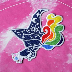 May include: Embroidered patch featuring a dark blue witch's hat with a crescent moon, stars, and an eye symbol. A rainbow-colored flame design emanates from the hat. The patch is on a pink background.