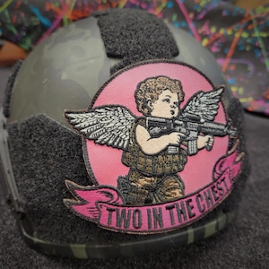 May include: A tactical helmet with a camouflage pattern and a patch featuring an angel holding a rifle. The patch is set on a pink background with the text "TWO IN THE CHEST".