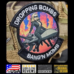 May include: A black embroidered patch with the text "DROPPING BOMBS" and "BANG'N MOMS". The patch features a cartoon woman in a black jumpsuit and sunglasses, sitting on a drone-like aircraft, with a fiery background. The patch is made in the USA.
