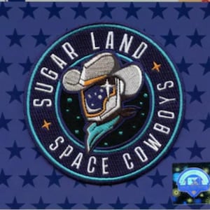 May include: Embroidered patch featuring a cowboy in a space helmet. The patch is circular with the words "Sugar Land Space Cowboys" in white lettering on a navy blue background. The background has a pattern of dark blue stars.