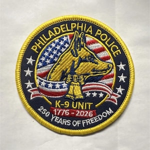 May include: Embroidered patch featuring the words "PHILADELPHIA POLICE" and "K-9 UNIT". The patch includes an image of a dog, the American flag, and the text "250 YEARS OF FREEDOM 1776-2026". The patch has a yellow border and a blue background.