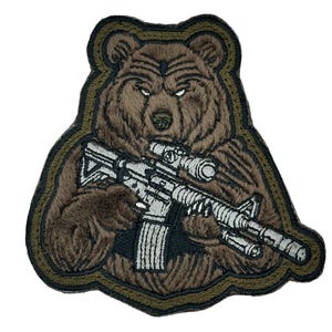 May include: Embroidered patch featuring a brown bear holding a silver rifle with a scope. The bear has a stern expression and is set against a dark green border. The patch has a textured, detailed design.