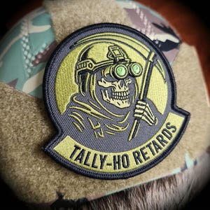 May include: A round patch featuring a skull wearing a helmet and night vision goggles, holding a scythe. The patch is embroidered in yellow and gray, with the words "TALLY-HO RETARDS" in yellow on a gray banner. The patch is attached to a camouflage fabric.