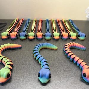Könnte beinhalten: Eine Sammlung farbenfroher, beweglicher Schlangenspielzeuge. Die Schlangen weisen einen Regenbogenfarbverlauf auf, darunter Blau, Grün, Orange und Gelb. Jede Schlange hat einen abgerundeten Kopf mit schwarzen Augen und einen segmentierten Körper.