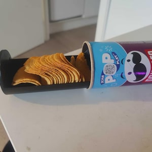 May include: A partially opened can of Pringles Texas BBQ Sauce chips with a black serving tray. The chips are golden brown and stacked. The can is blue and purple with the Pringles logo and the words "SCAN POP PLAY".