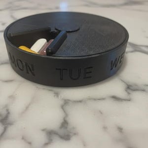 May include: A black, round pill organizer with the days of the week labeled. The lid is partially open, revealing several pills in various colors. The organizer is sitting on a marble surface.