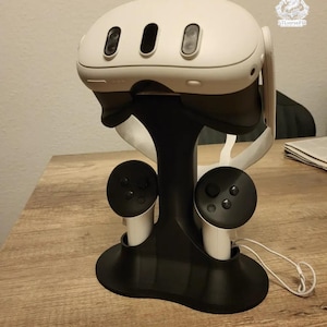 Op de afbeelding: Een witte en zwarte VR-headset en controllerstandaard. De standaard houdt de VR-headset en twee controllers vast. De standaard is zwart met witte accenten. De VR-headset heeft drie lenzen aan de voorkant.
