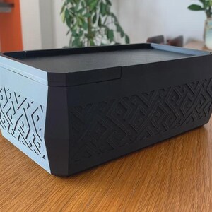 May include: A rectangular storage box with a two-tone design. The top and sides are black, while the front and back are light gray. The sides feature a geometric pattern. The box is on a wooden table.