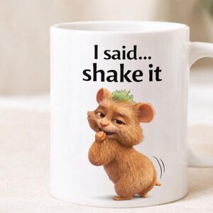 May include: White ceramic mug with a cartoon illustration of a brown furry creature with a green tuft of hair. The mug has the text "I said... shake it" in black. The mug has a curved handle.