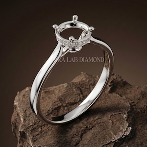 May include: A silver-toned ring setting featuring a four-prong head designed to hold a center stone. The gallery is accented with a row of small, clear pavé-set stones. The text AURA LAB DIAMOND is visible on the band of this polished metal ring.