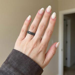 May include: A black ring set with small, dark gemstones. The ring is worn on a hand with long, oval-shaped nails. The image includes the text "AURA LAB DIAMOND".