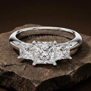 Pode incluir: Anel de noivado prateado com três diamantes de corte quadrado. O anel tem uma banda polida e o texto "AURA LAB DIAMOND" está gravado no anel. O anel está sobre uma superfície de pedra.