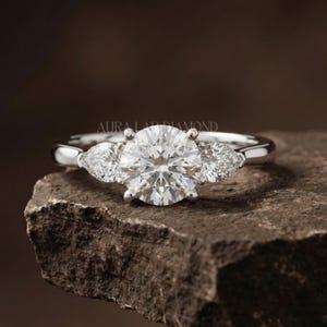 May include: A platinum engagement ring featuring a large, round-cut diamond and two pear-shaped side stones. The ring is displayed on a dark, textured surface. The brand name "AURA LAB DIAMOND" is visible.