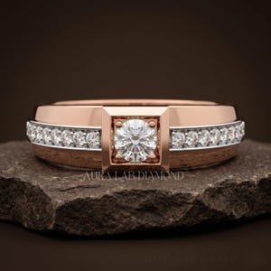 May include: A rose gold ring featuring a central round brilliant-cut diamond set in a square frame, flanked by a channel of smaller round diamonds along the band. The text AURA LAB DIAMOND is visible on the surface below the ring.