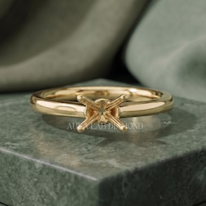 May include: A polished yellow gold ring setting featuring a four-prong head designed to hold a single gemstone. The band is smooth and rounded, with the text AURA LAB DIAMOND engraved on the metal surface near the centre prong setting.