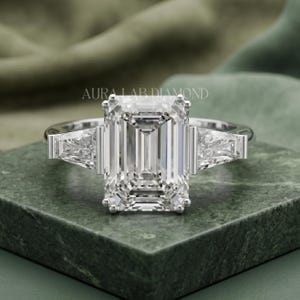 May include: A silver-coloured ring featuring a large emerald-cut centre stone flanked by two smaller trapezoid-cut side stones. The band is polished metal. The text AURA LAB DIAMOND is visible above the ring, which is set on a green surface.