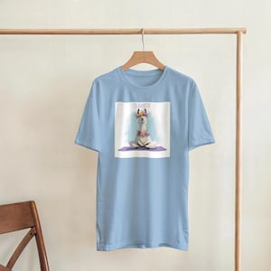 Op de afbeelding: Lichtblauw T-shirt met een afbeelding van een lama in een yogahouding, versierd met een bloemenkrans en ketting. De tekst "LLAMASTE" staat boven de lama. Het T-shirt hangt aan een houten kledingrek.