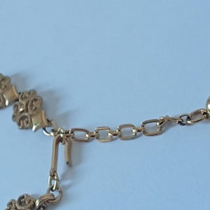 May include: Close-up of a gold-colored necklace. The necklace features ornate floral-like designs and a chain with a small ball at the end. The clasp is visible, and the background is a light blue.