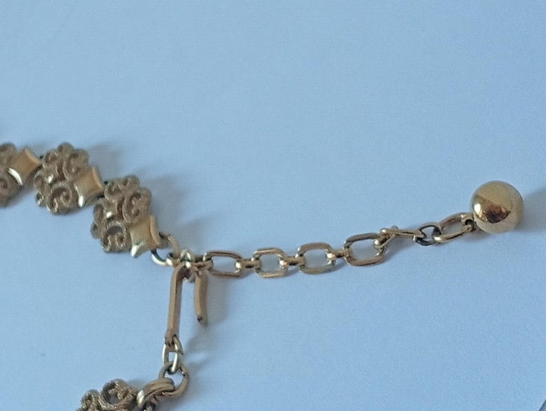 May include: Close-up of a gold-colored necklace. The necklace features ornate floral-like designs and a chain with a small ball at the end. The clasp is visible, and the background is a light blue.
