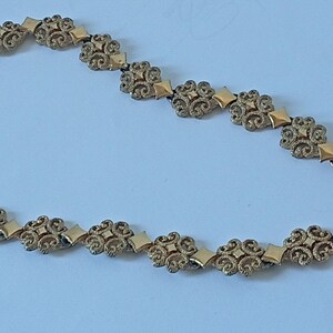 May include: A gold-toned necklace with an ornate floral and geometric design. The necklace features repeating floral elements connected by diamond-shaped links. The clasp is a simple hook and eye closure. The jewelry is set against a light blue background.