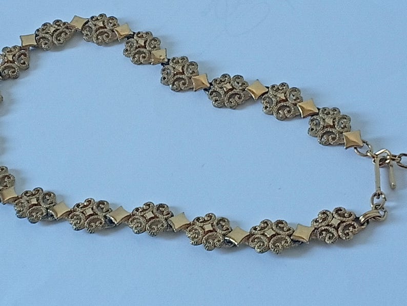 May include: A gold-toned necklace with an ornate floral and geometric design. The necklace features repeating floral elements connected by diamond-shaped links. The clasp is a simple hook and eye closure. The jewelry is set against a light blue background.