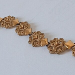 May include: A gold-toned necklace featuring ornate floral and diamond-shaped elements. The necklace is composed of multiple decorative pieces connected by a dark cord, creating a vintage or antique aesthetic.