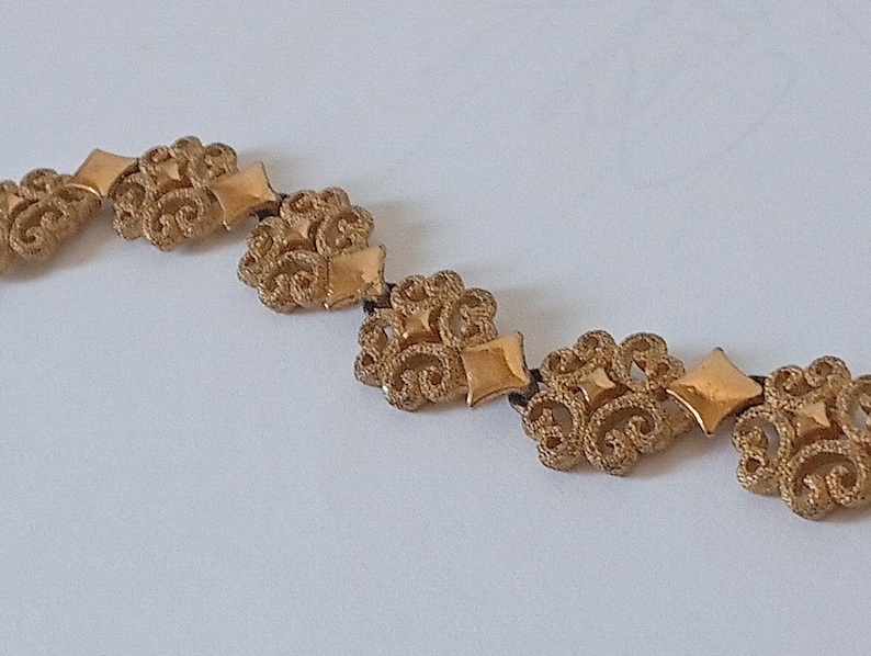 May include: A gold-toned necklace featuring ornate floral and diamond-shaped elements. The necklace is composed of multiple decorative pieces connected by a dark cord, creating a vintage or antique aesthetic.