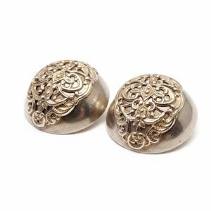 Puede incluir: Dos pendientes plateados en forma de cúpula. La parte superior de cada pendiente presenta un diseño calado ornamentado con motivos florales y de volutas. Los pendientes tienen una superficie pulida y reflectante sobre fondo blanco.