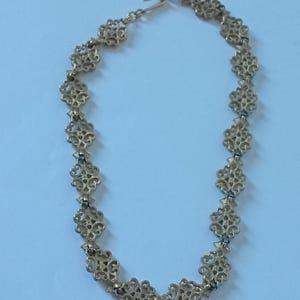 May include: Gold-toned necklace featuring an openwork design with repeating floral-like motifs. The necklace has a delicate chain and a hook clasp. The intricate pattern and warm color suggest a vintage or antique style.