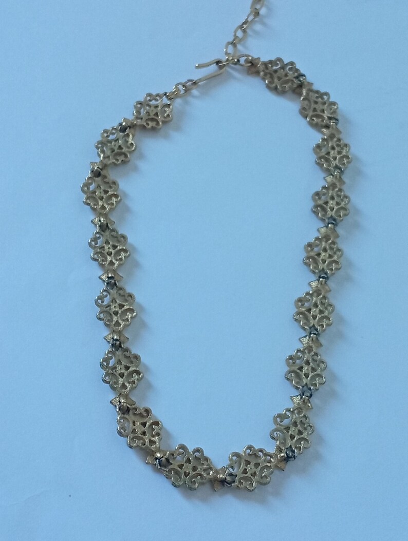 May include: Gold-toned necklace featuring an openwork design with repeating floral-like motifs. The necklace has a delicate chain and a hook clasp. The intricate pattern and warm color suggest a vintage or antique style.