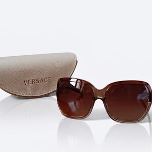 May include: Brown-tinted sunglasses with a matching case. The sunglasses have a rounded, oversized frame. The case is a light beige colour and has the word "VERSACE" printed on it.