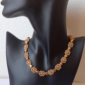 May include: Gold-toned necklace with an ornate, openwork design. The necklace features repeating floral and diamond-shaped elements, displayed on a black mannequin. The jewelry is a classic accessory.