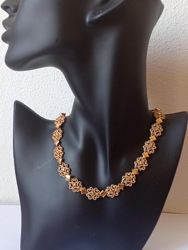 May include: Gold-toned necklace with an ornate, openwork design. The necklace features repeating floral and diamond-shaped elements, displayed on a black mannequin. The jewelry is a classic accessory.